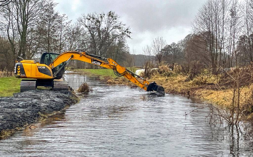 Koparka gąsienicowa JCB wykonująca odmulanianie koryta rzeki z brzegu, w trudnych warunkach terenowych.