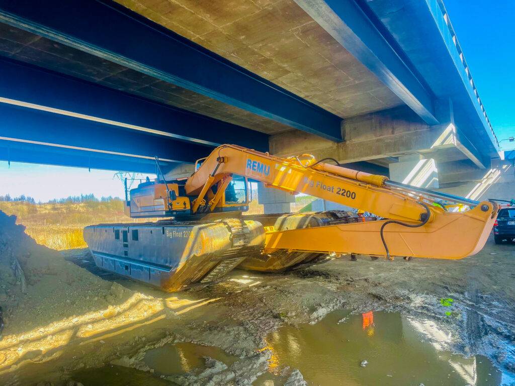 Koparka pływająca REMU Big Float 2200 pracująca pod betonowym mostem na grząskim i zalanym terenie.