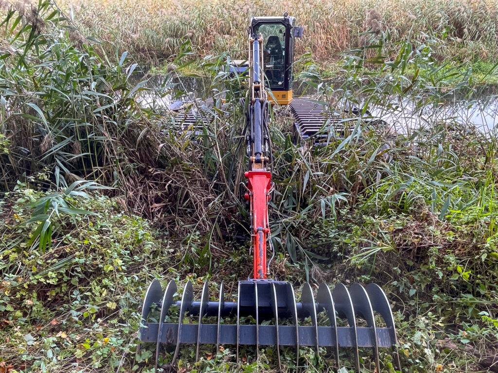 Koparka pływająca z chwytakiem pracująca wśród trzcin nad brzegiem zbiornika wodnego.